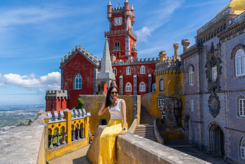 Shivani posing in front of the colorful Pena Palace in Sintra, Portugal, wearing a white and yellow outfit against the red and yellow palace